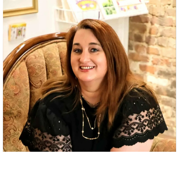 Coach Cinnamon B with red hair smiling, wearing black lace top, sitting on a patterned chair.