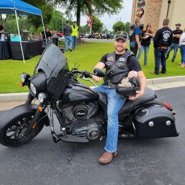 Man sitting on a black motorcycle holding a helmet, smiling.