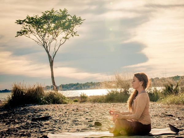 A woman meditating on a beach during sunset with a tree nearby.