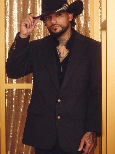 Man in a black blazer and cowboy hat poses against a glittering gold backdrop.