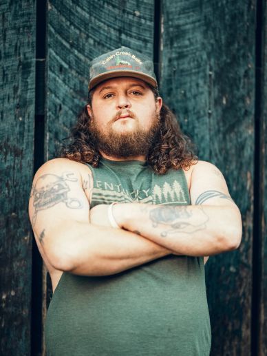 Man with long curly hair, beard, and a cap standing against a wooden background.