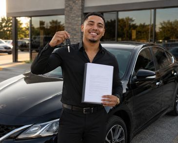Man joyfully holding car keys and paperwork beside a black car.