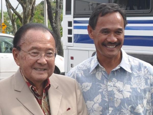 Two men smiling outdoors, one in a beige suit and the other in a floral shirt.