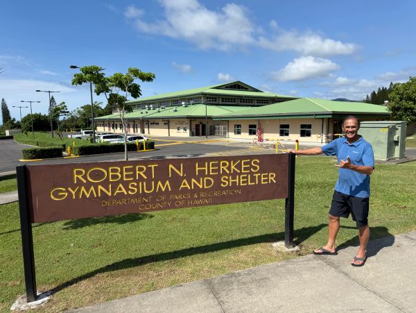 Man posing next to Robert N. Herkes Gymnasium and Shelter sign under a sunny sky.