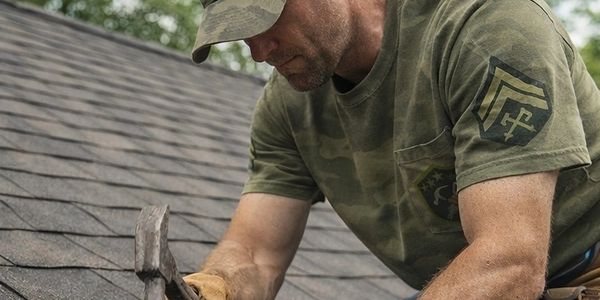 Man in camouflage shirt and hat working on shingled roof with hammer.