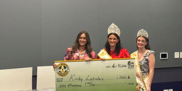 Three women, two wearing crowns, holding a large check for $1,000.
