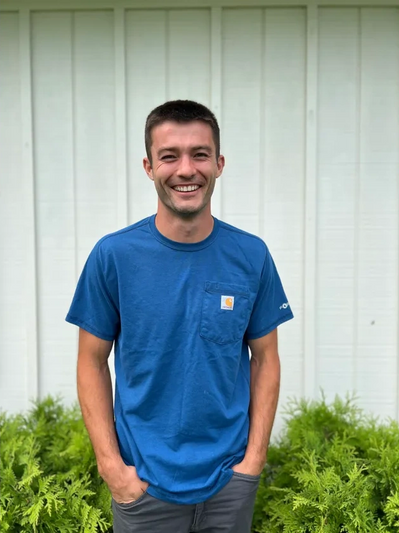 A smiling man stands with hands in pockets, wearing a blue shirt in front of a white wall and green plants.