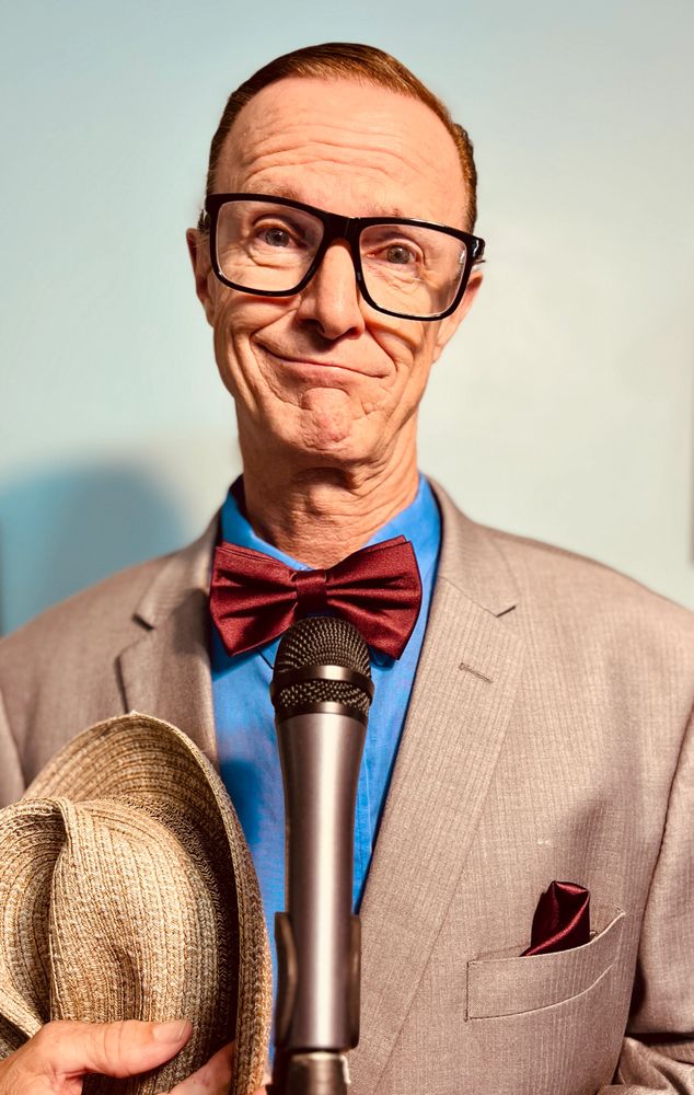 Man in a suit with bow tie holding a hat and standing behind a microphone.