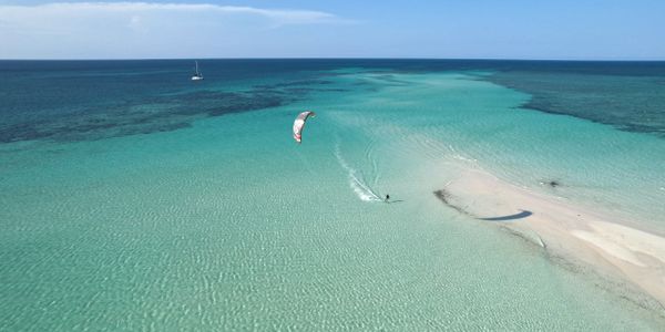 Kite surfer glides over clear turquoise waters near a sandy sandbar under a sunny sky.