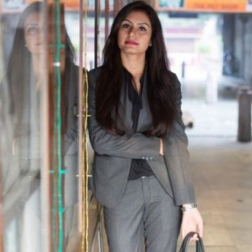 Confident woman in a gray suit leaning against a glass wall, holding a handbag.