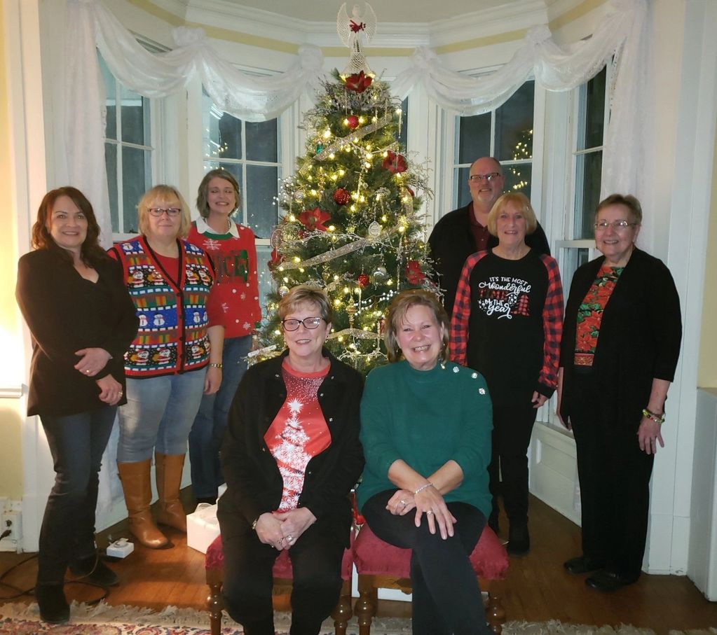 A group of eight adults posing cheerfully by a decorated Christmas tree indoors.