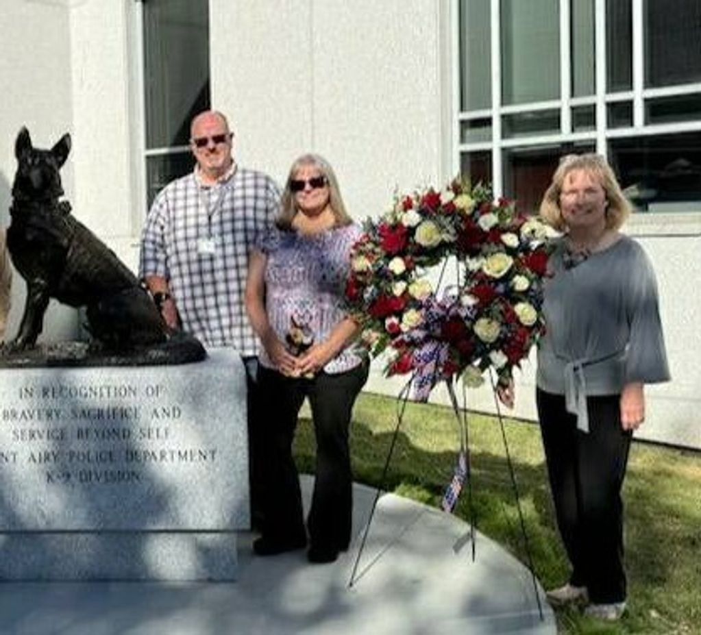 Three people stand beside a police dog statue and a floral wreath outdoors.