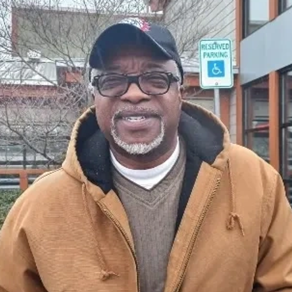 Smiling man in glasses and brown jacket outdoors.