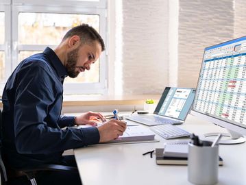 A man working at a desk with spreadsheets on computer screens.