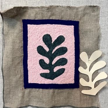 Fabric embroidery of a green leaf design on pink and blue background.
