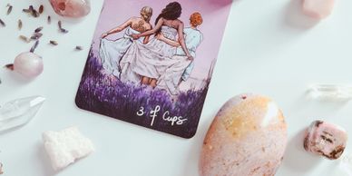 Tarot card 3 of Cups surrounded by crystals on a white surface.