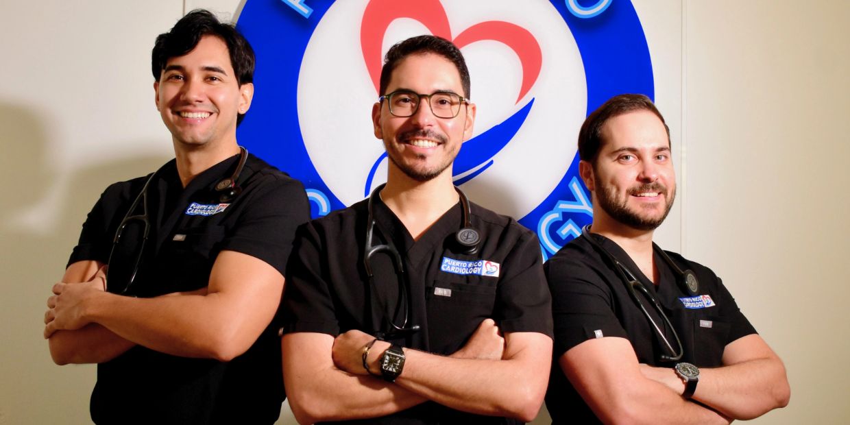 Three smiling doctors in black scrubs with stethoscopes, standing confidently in front of a cardiology sign.