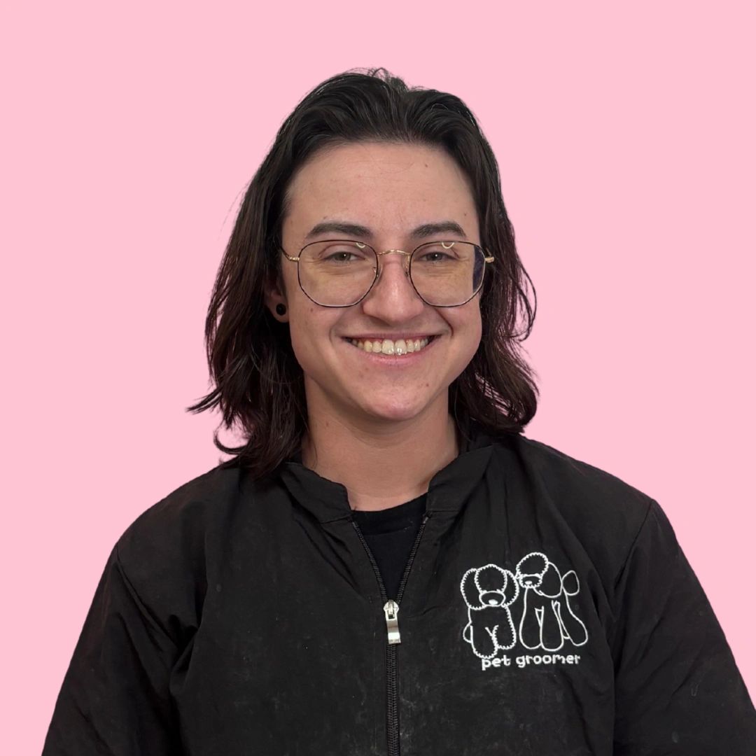 Smiling person with glasses wearing a pet groomer jacket against a pink background.