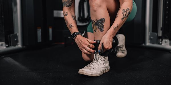Person with tattoos tying worn white sneakers in a gym setting.