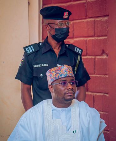 A man in traditional attire seated with a police officer standing behind.