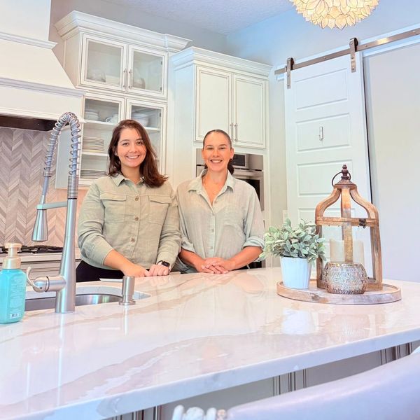 Two women smiling in a bright modern kitchen with floral pendant lights.