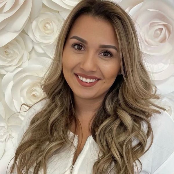 A smiling woman with long wavy hair in front of a white floral background.