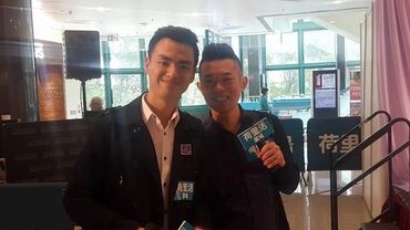Two men posing indoors at a formal event, holding booklets.