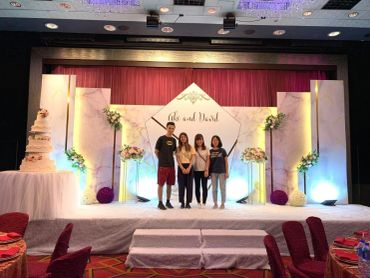 Four people standing on a decorated stage with floral arrangements and a wedding cake.