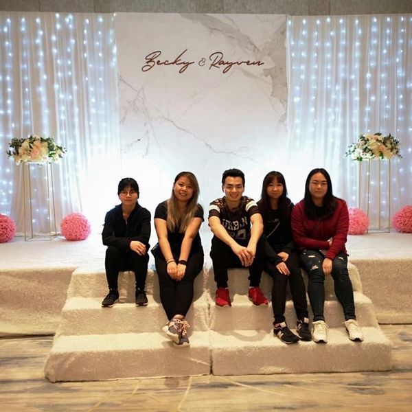 Five friends sit on white steps in front of a decorated backdrop with lights and flowers.