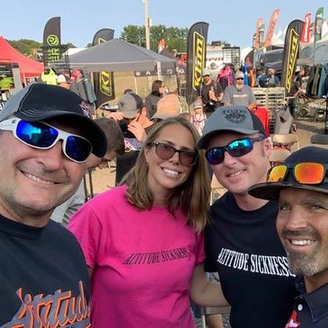 Four friends smiling at an outdoor event, wearing sunglasses and casual clothes.