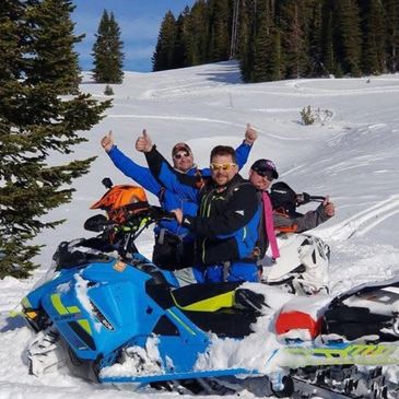 Three people on snowmobiles giving thumbs up in a snowy forest.
