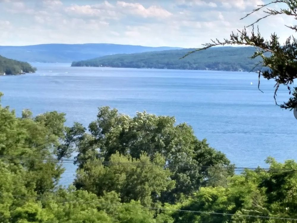 Serene lake view surrounded by lush green trees and distant hills under a partly cloudy sky.