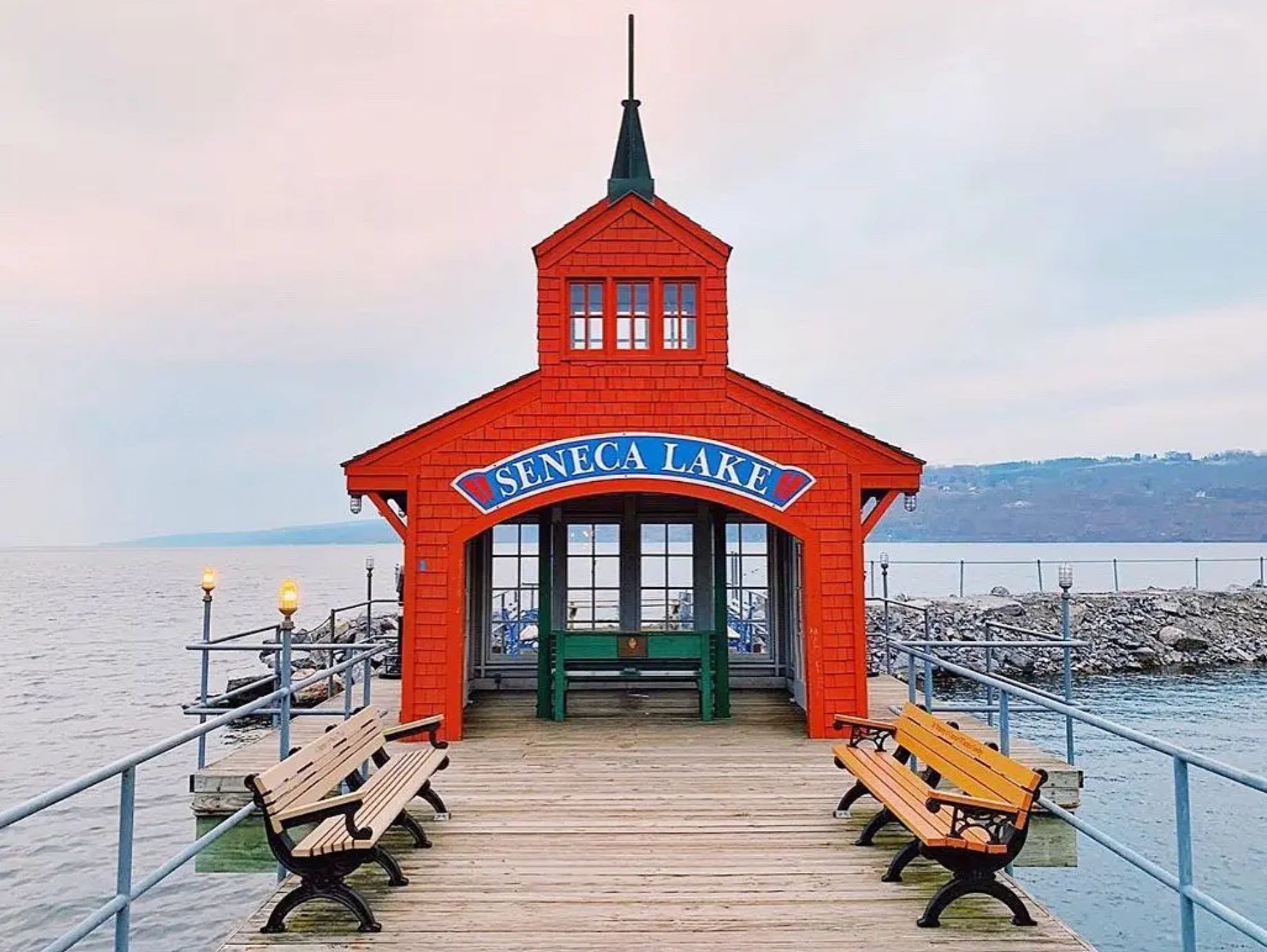 Red pavilion with 'Seneca Lake' sign on a pier surrounded by benches and water.