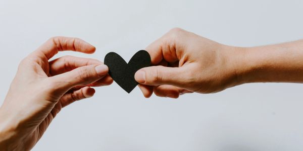 Two hands exchanging a small black paper heart against a plain background.