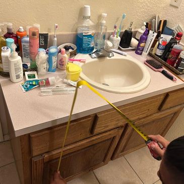 Bathroom countertop cluttered with toiletries and a person measuring cabinet width.