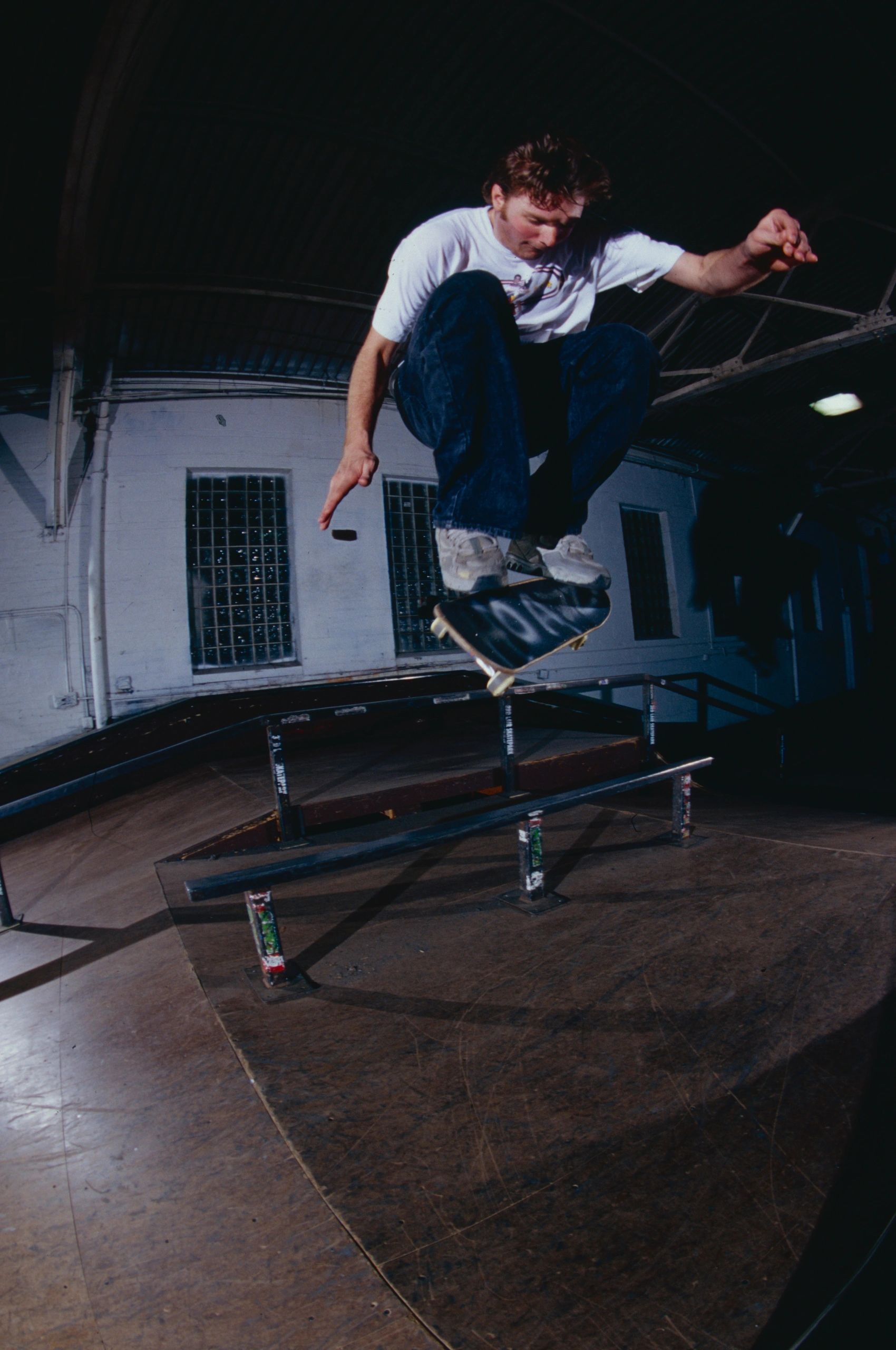 Skateboarder performing a trick over a rail in an indoor skatepark.