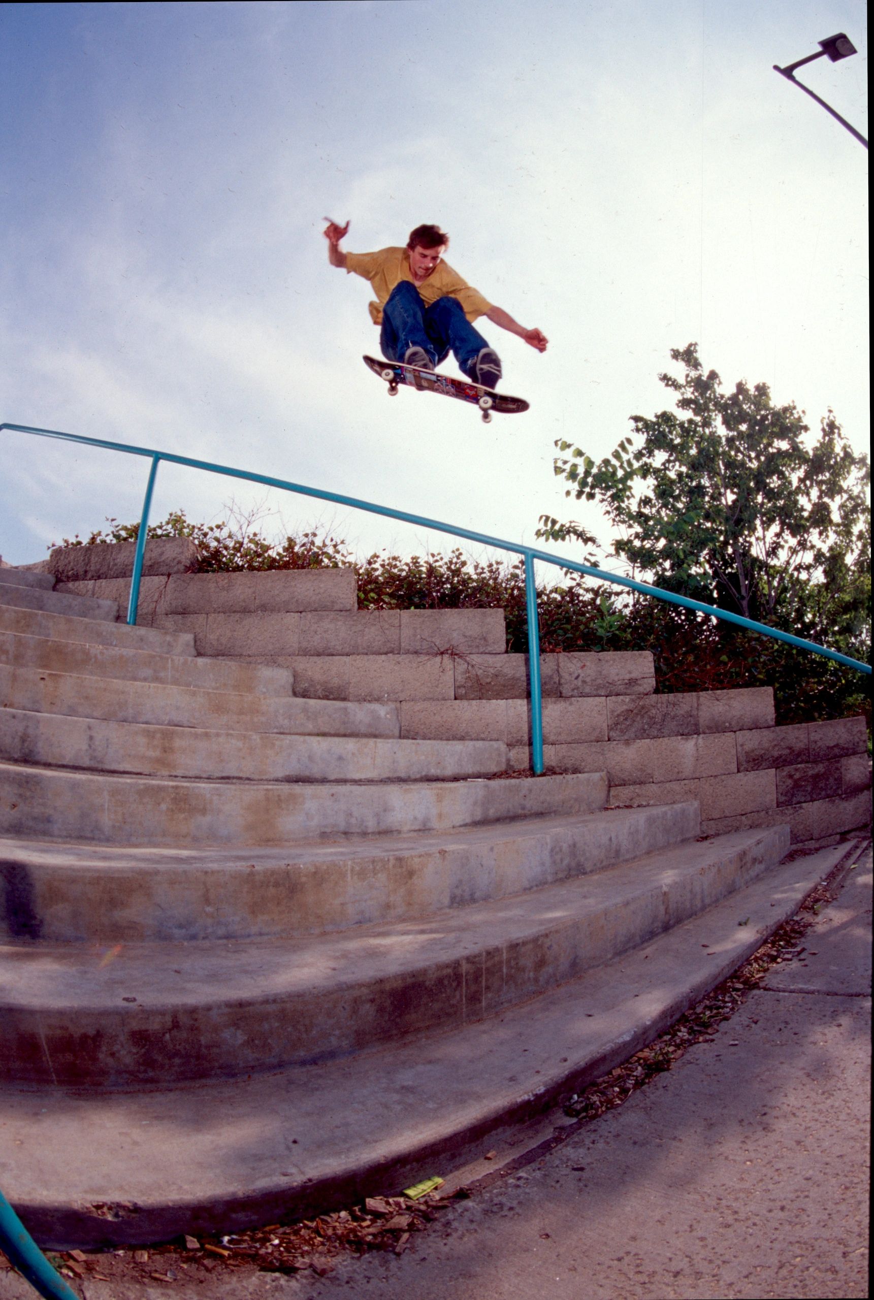 Skateboarder performing an ollie over a long stair rail outdoors.