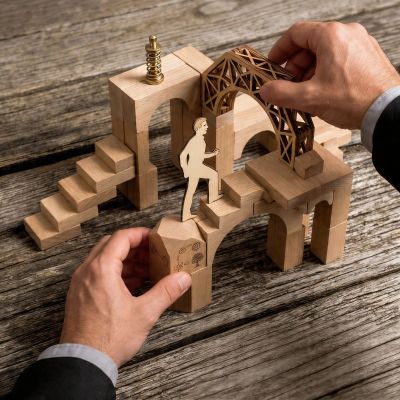 Hands assembling a wooden architectural puzzle with a paper figure walking on it.