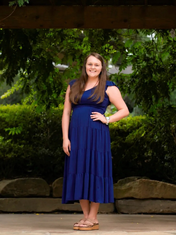 Woman in a blue dress standing outdoors under greenery.