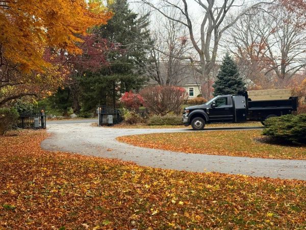 Black pickup truck parked by a driveway surrounded by autumn trees and fallen leaves.