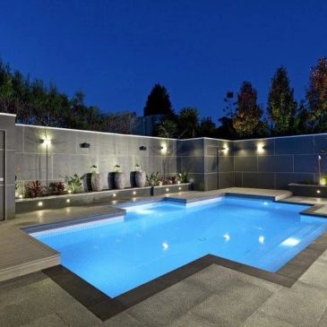 Modern backyard pool illuminated at night with water features and seating.
