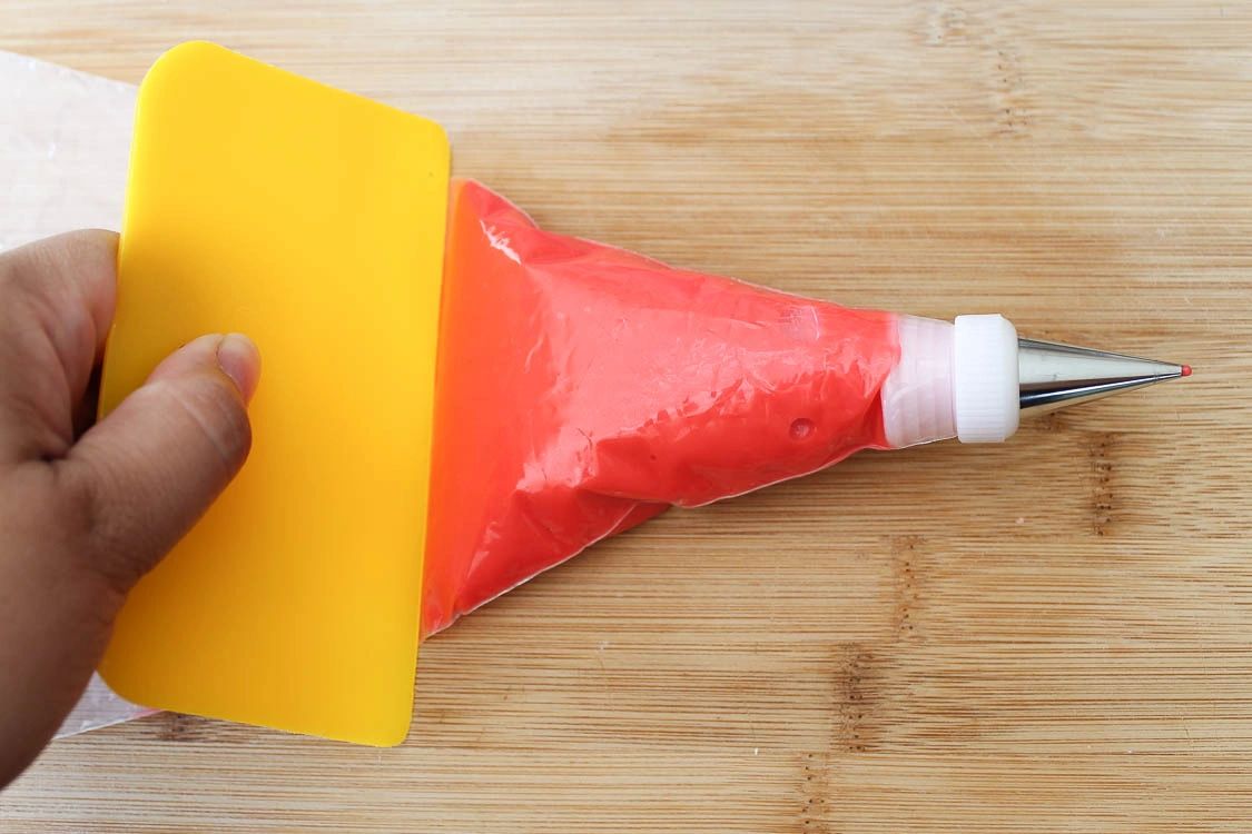 Red royal icing in piping bag with decorating tip being pushed with scraper.