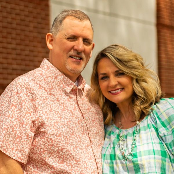 A smiling couple stands close together outside against a brick building.