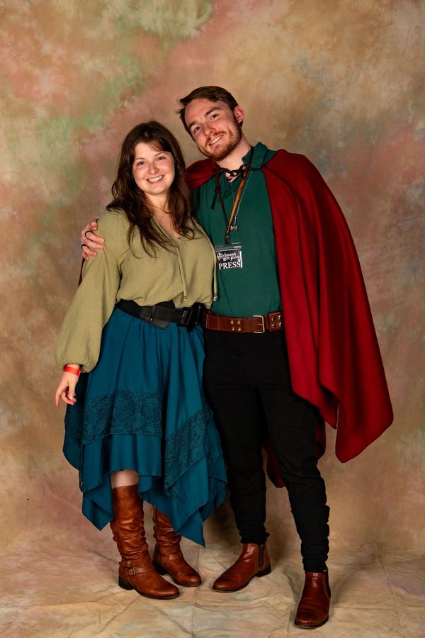 A smiling couple in medieval-style attire posing together against a soft backdrop.