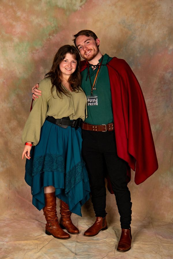 A smiling couple in medieval-style clothing posing together against a neutral backdrop.