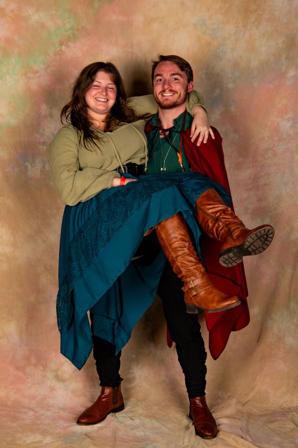 A man carries a smiling woman in a blue skirt and brown boots against a warm backdrop.