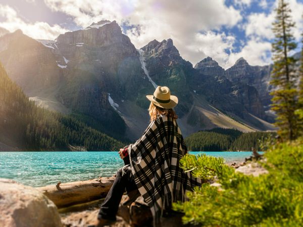 Person in a hat and plaid shawl gazing at turquoise lake and mountain range.