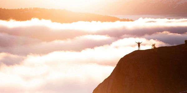 Person standing triumphantly on a mountain peak above clouds at sunrise.