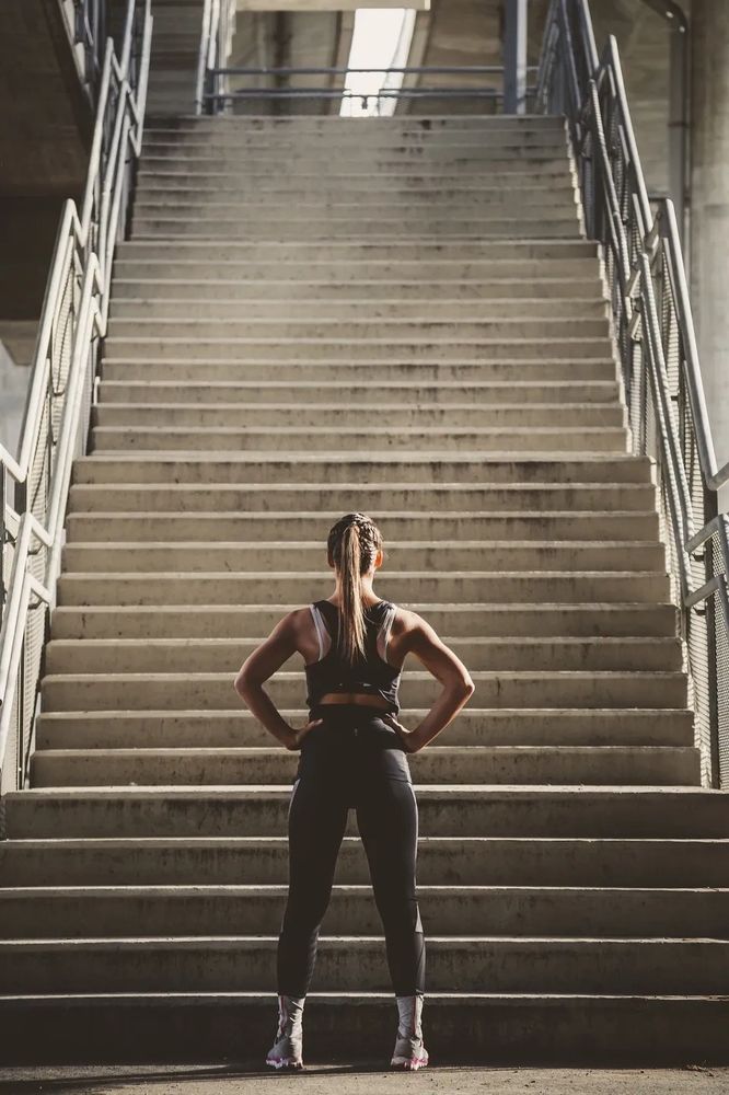 Woman in workout clothing, standing at the bottom of a tall staircase.