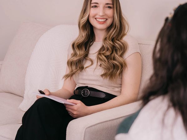 A woman with long wavy hair smiles while holding a notepad during a conversation.